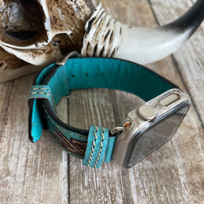 A turquoise leather watch band with brown stitched detailing and a silver metal watch case, displayed on a rustic wooden surface next to a horned skull prop.