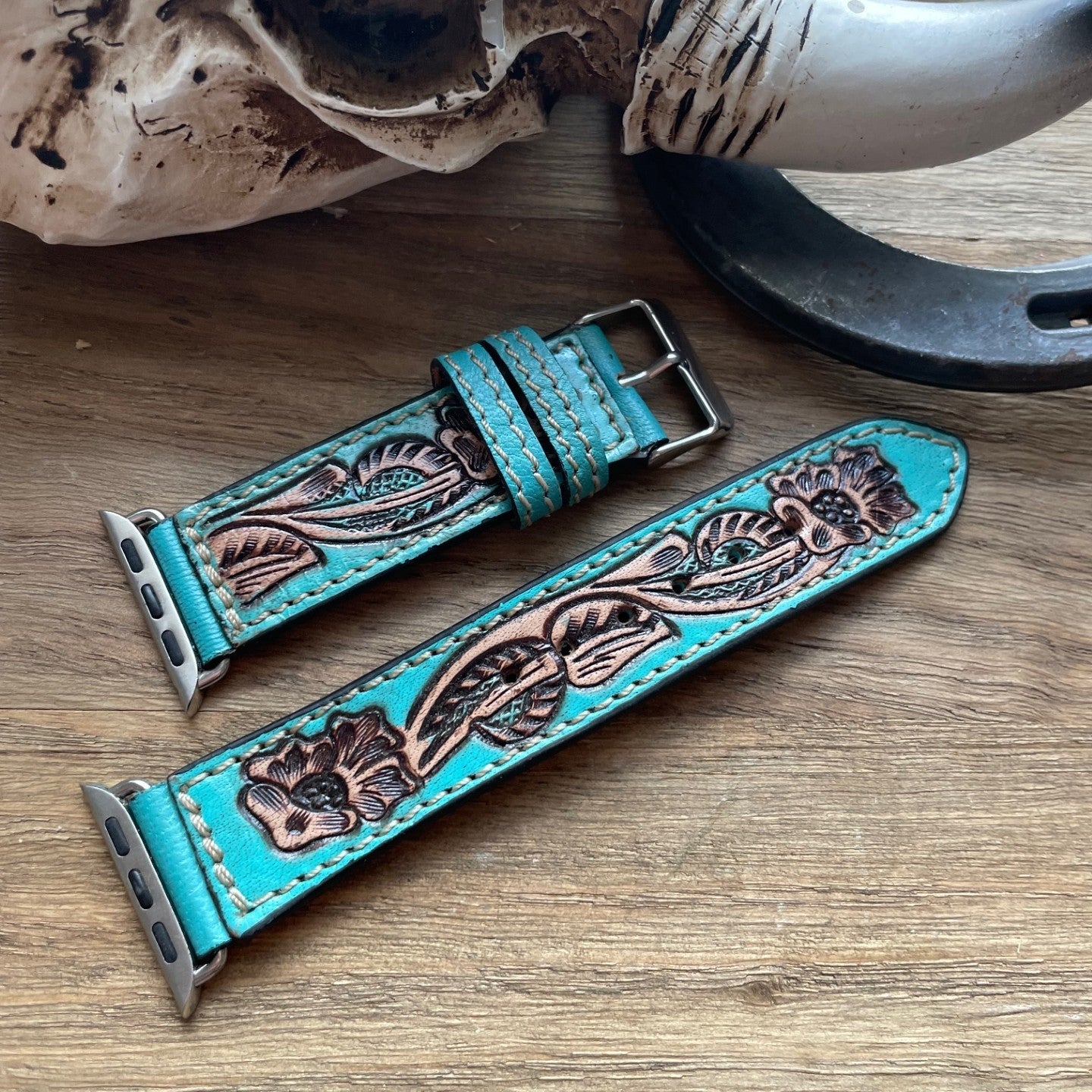 A turquoise leather watch strap with hand-tooled floral detailing and silver-toned hardware, laid on a wooden surface with rustic props including a horn, skull, and horseshoe.