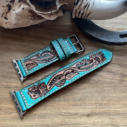 A turquoise leather watch strap with hand-tooled floral detailing and silver-toned hardware, laid on a wooden surface with rustic props including a horn, skull, and horseshoe.