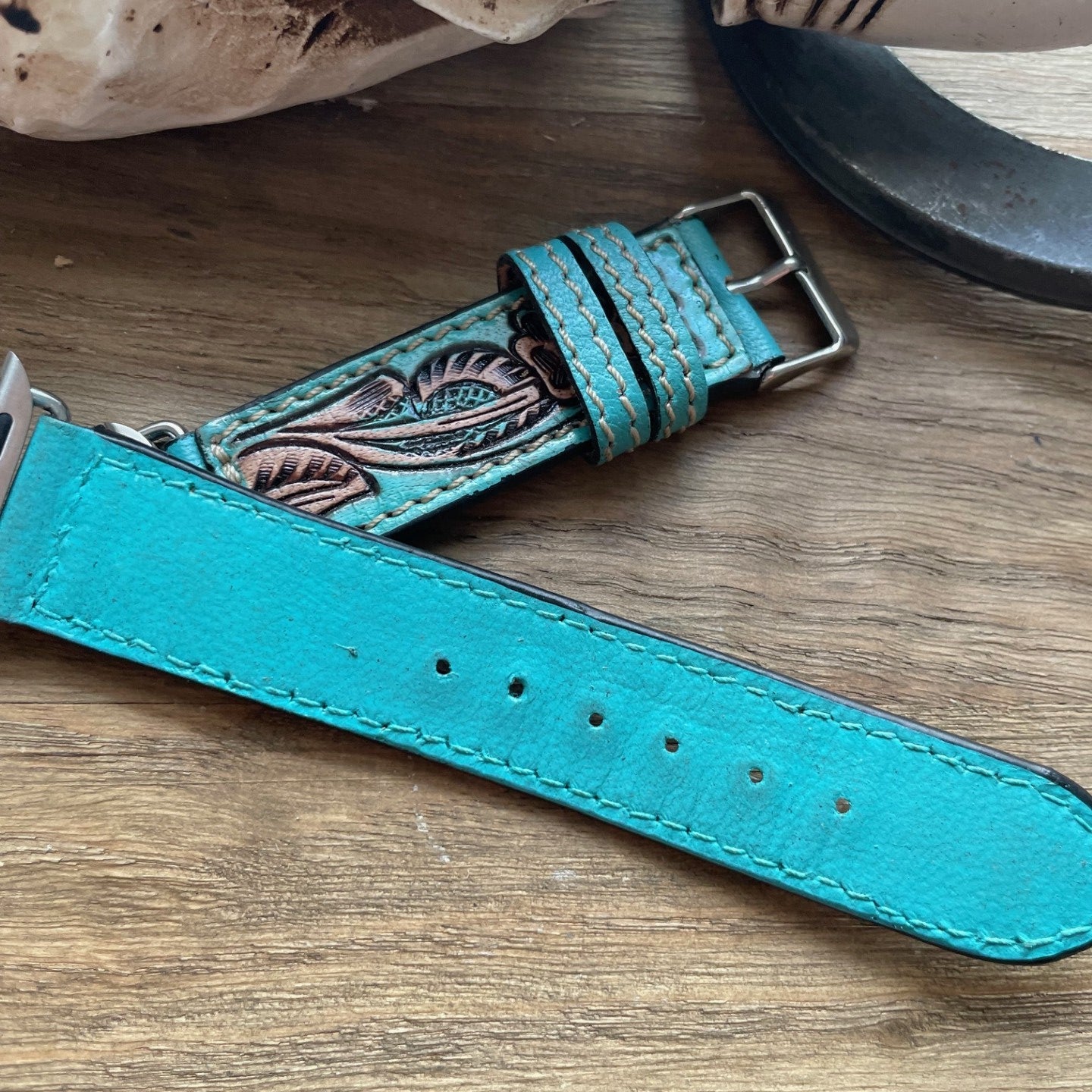 A turquoise leather watch strap with hand-tooled floral detailing, displayed on a rustic wooden surface next to a horn and bone prop.