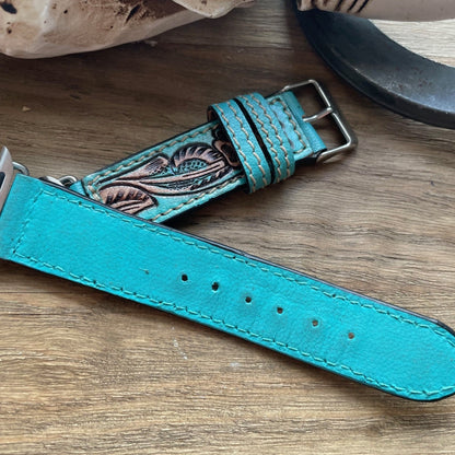 A turquoise leather watch strap with hand-tooled floral detailing, displayed on a rustic wooden surface next to a horn and bone prop.