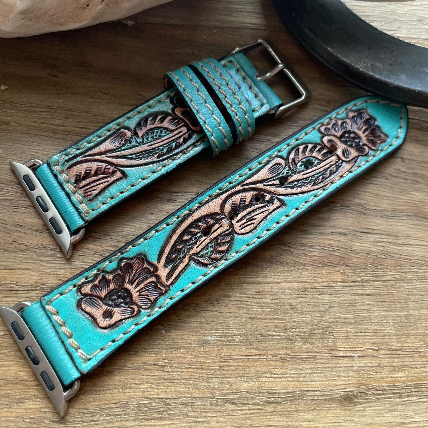 A turquoise leather watch strap with hand-tooled floral and leaf patterns, displayed on a wooden surface next to a ceramic object.