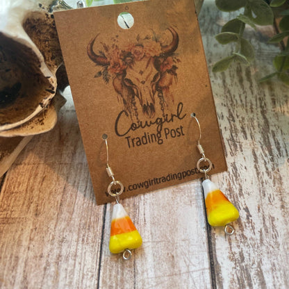 A pair of dangling earrings with candy corn beads, hanging from a brown paper display card printed with 'Cowgirl Trading Post'. The card is styled on a rustic wooden surface next to a skull and green foliage.