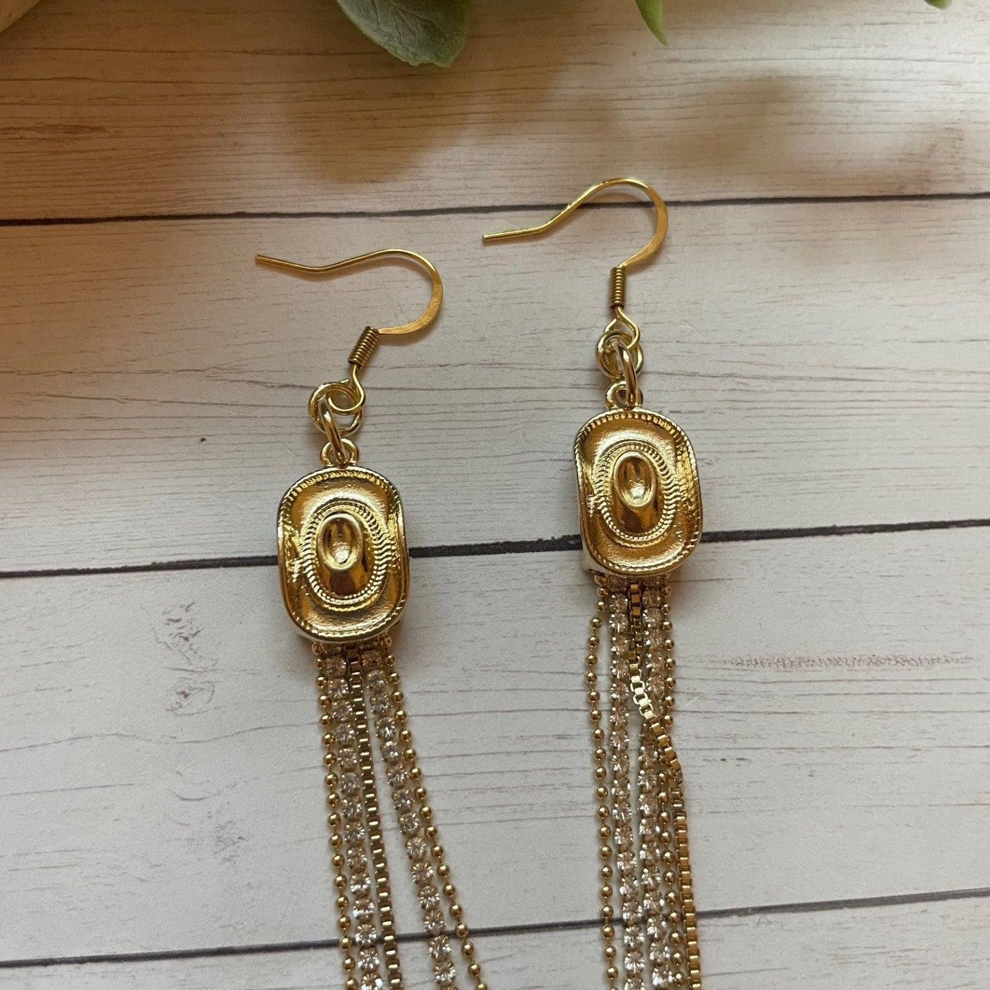 Gold tassel earrings on a wooden surface with green leaves in the background