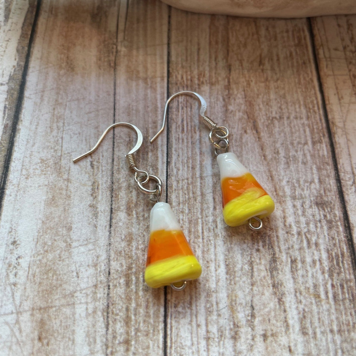 Candy corn dangle earrings with silver fishhook wires, featuring white, orange, and yellow striped beads on a rustic wooden surface.