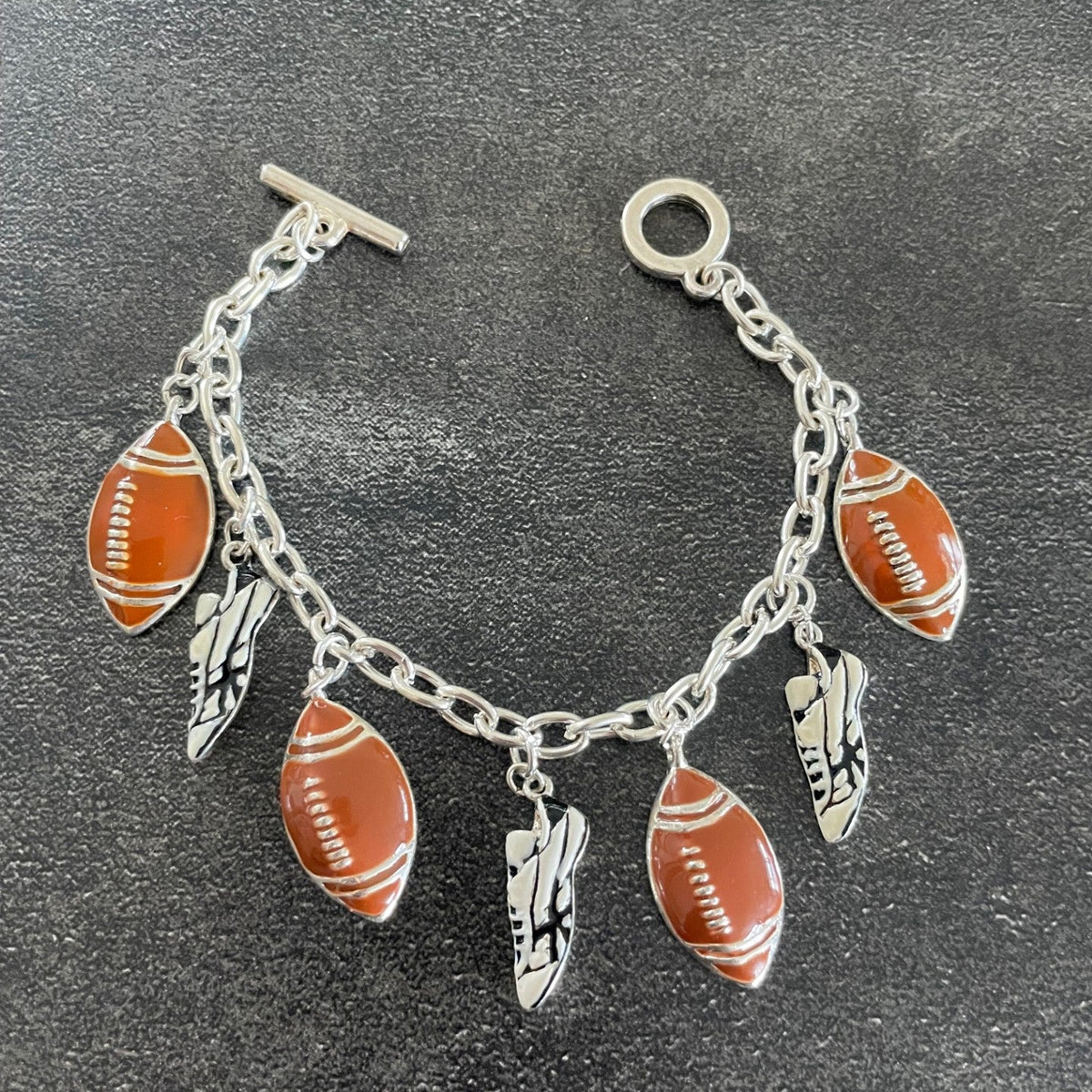 A silver-tone chain bracelet with football-shaped charms, feather charms, and shoe charms, displayed on a dark textured surface.