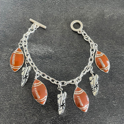 A silver-tone chain bracelet with football-shaped charms, feather charms, and shoe charms, displayed on a dark textured surface.