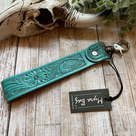 Turquoise leather keychain with intricate designs on a wooden surface with a skull and plant in the background.
