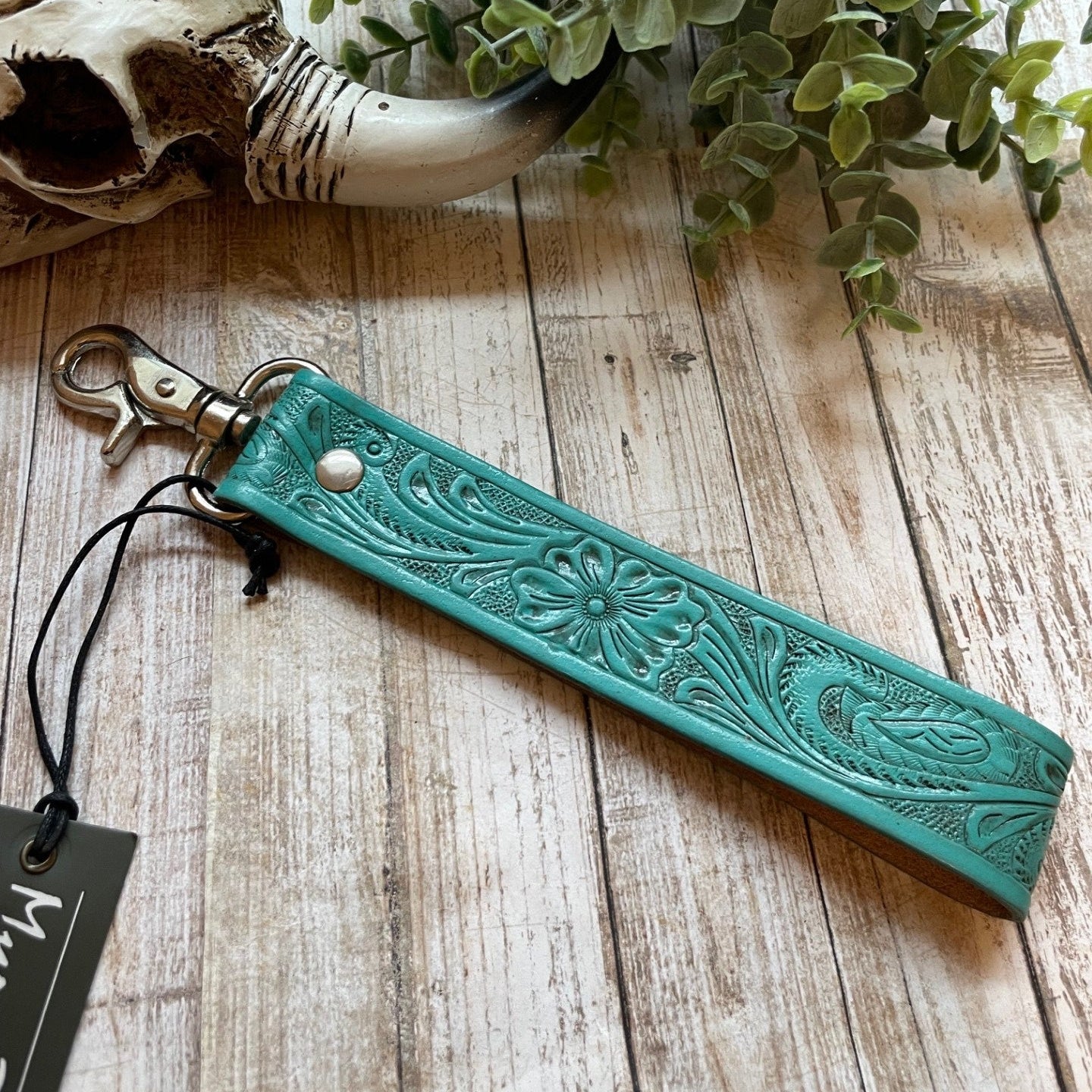 Turquoise leather strap with floral patterns on a wooden surface with a plant and skull decoration.
