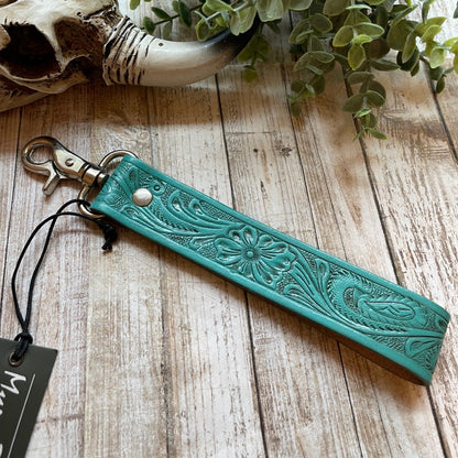 Turquoise leather strap with floral patterns on a wooden surface with a plant and skull decoration.