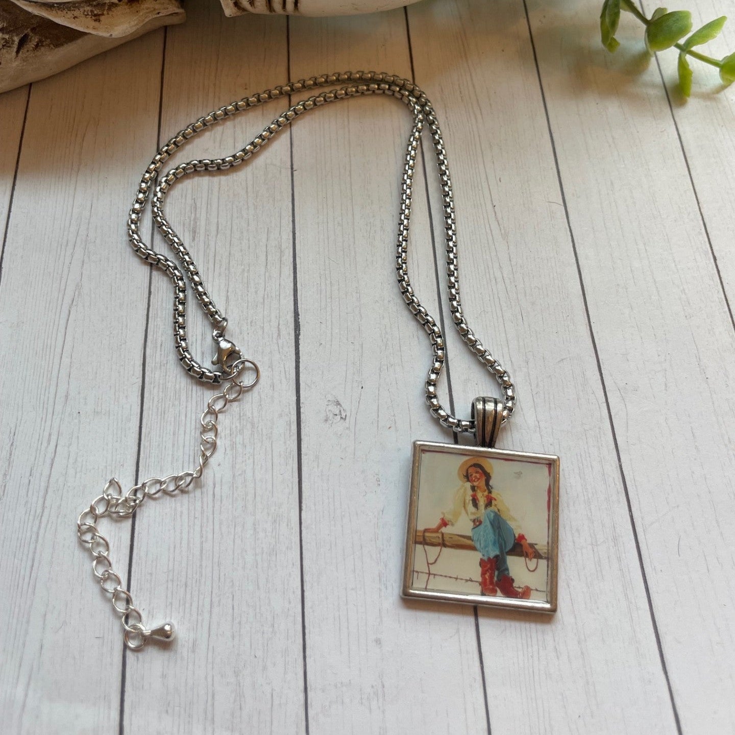 Necklace with a pendant featuring a girl in a blue dress on a wooden surface with a skull and plant in the background.