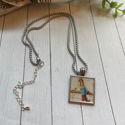 Necklace with a pendant featuring a girl in a blue dress on a wooden surface with a skull and plant in the background.