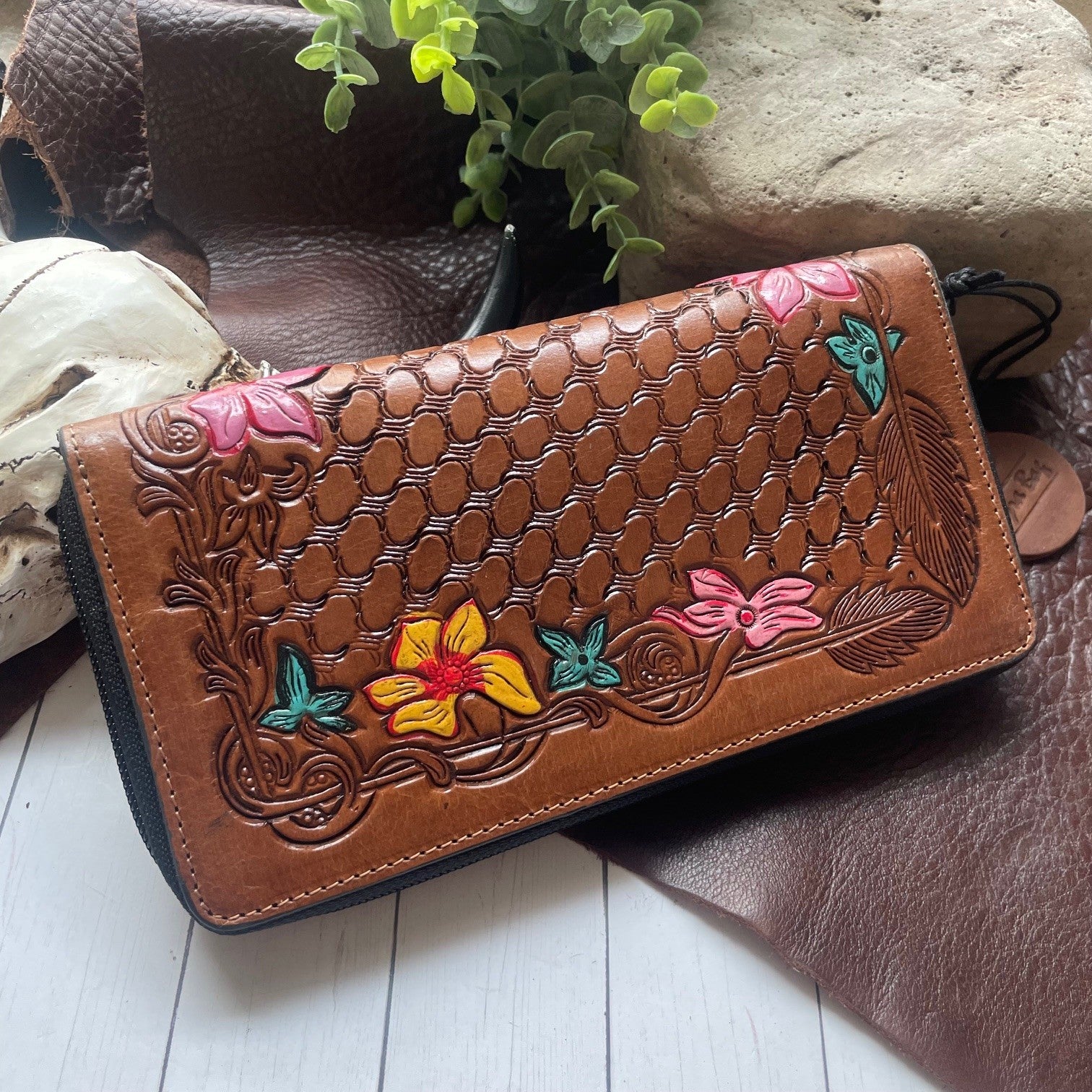 A brown tooled leather wallet with floral and butterfly designs, displayed on a rustic surface with leather and a plant in the background.
