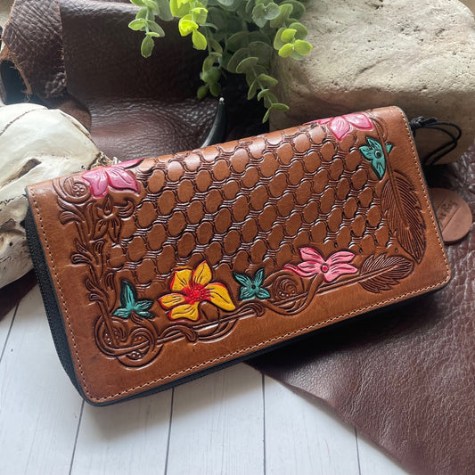 A brown tooled leather wallet with floral and butterfly designs, displayed on a rustic surface with leather and a plant in the background.