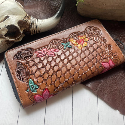 A brown tooled leather wallet with floral and dragonfly embossing, displayed on a white wood surface with a skull prop and dark brown leather background.