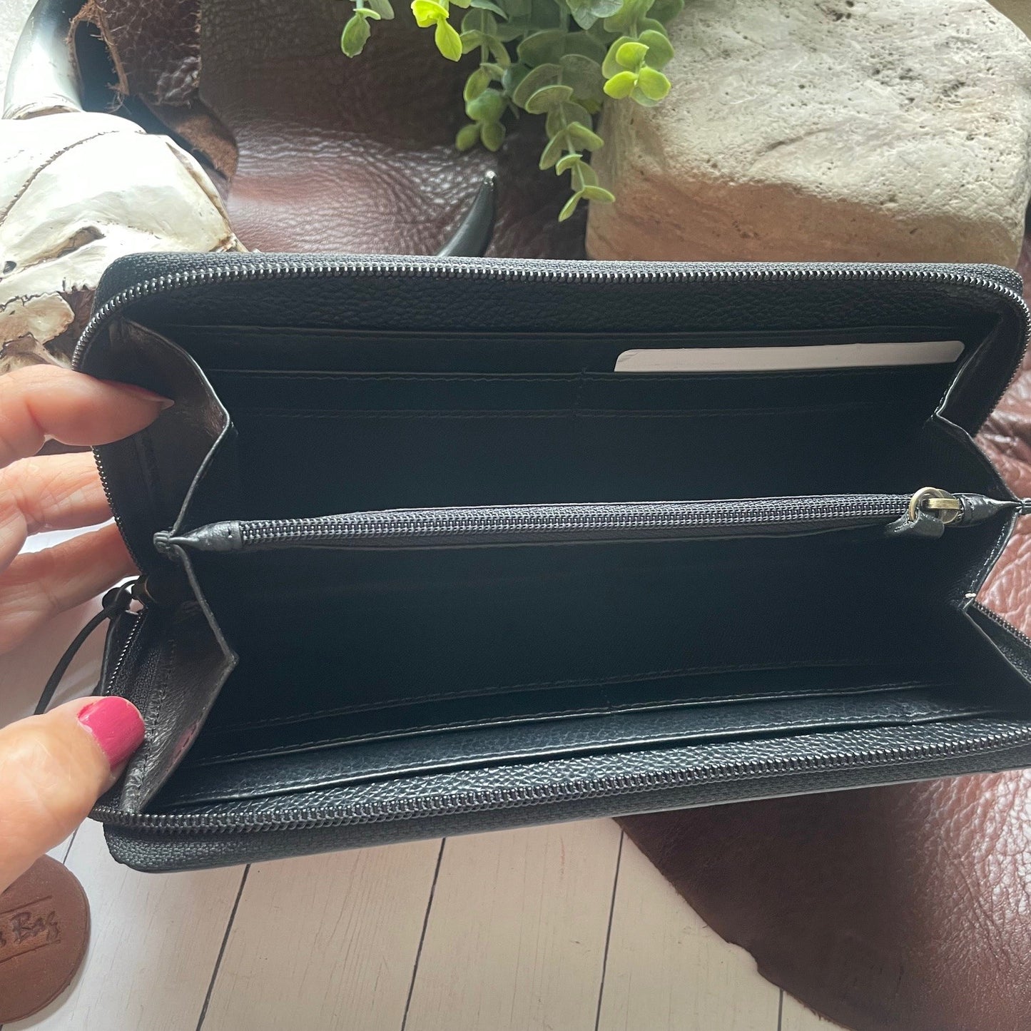A black textured leather zip-around wallet held open by a hand with pink nail polish, showing multiple interior card slots and a horizontal zip compartment. The wallet is photographed over a brown leather surface with a plant and stone in the background.