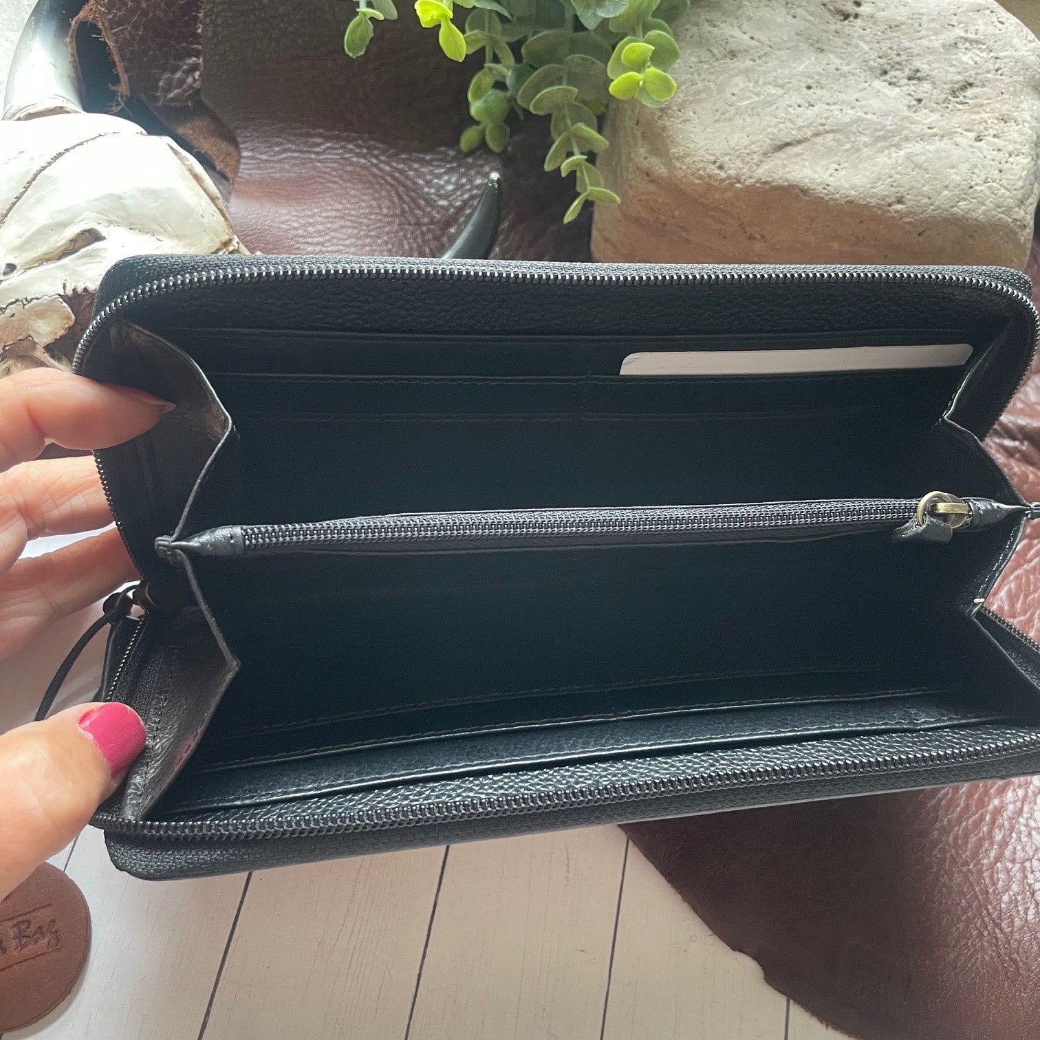 A black textured leather zip-around wallet held open by a hand with pink nail polish, showing multiple interior card slots and a horizontal zip compartment. The wallet is photographed over a brown leather surface with a plant and stone in the background.