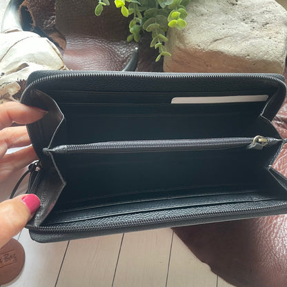 A black textured leather zip-around wallet held open by a hand with pink nail polish, showing multiple interior card slots and a horizontal zip compartment. The wallet is photographed over a brown leather surface with a plant and stone in the background.