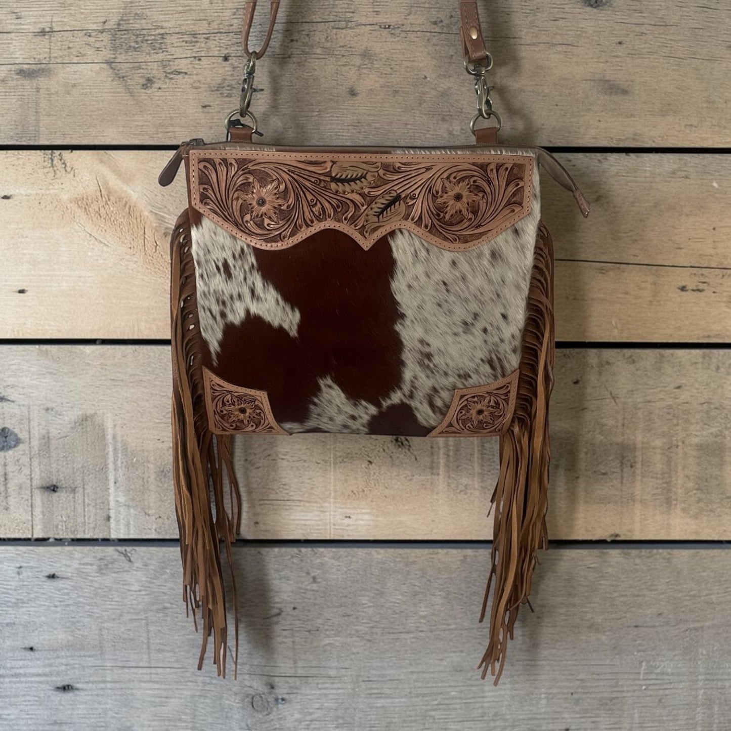 A western-style crossbody bag with a brown and white cowhide body, tooled leather trim, and long fringe, hanging against a rustic wood plank background.