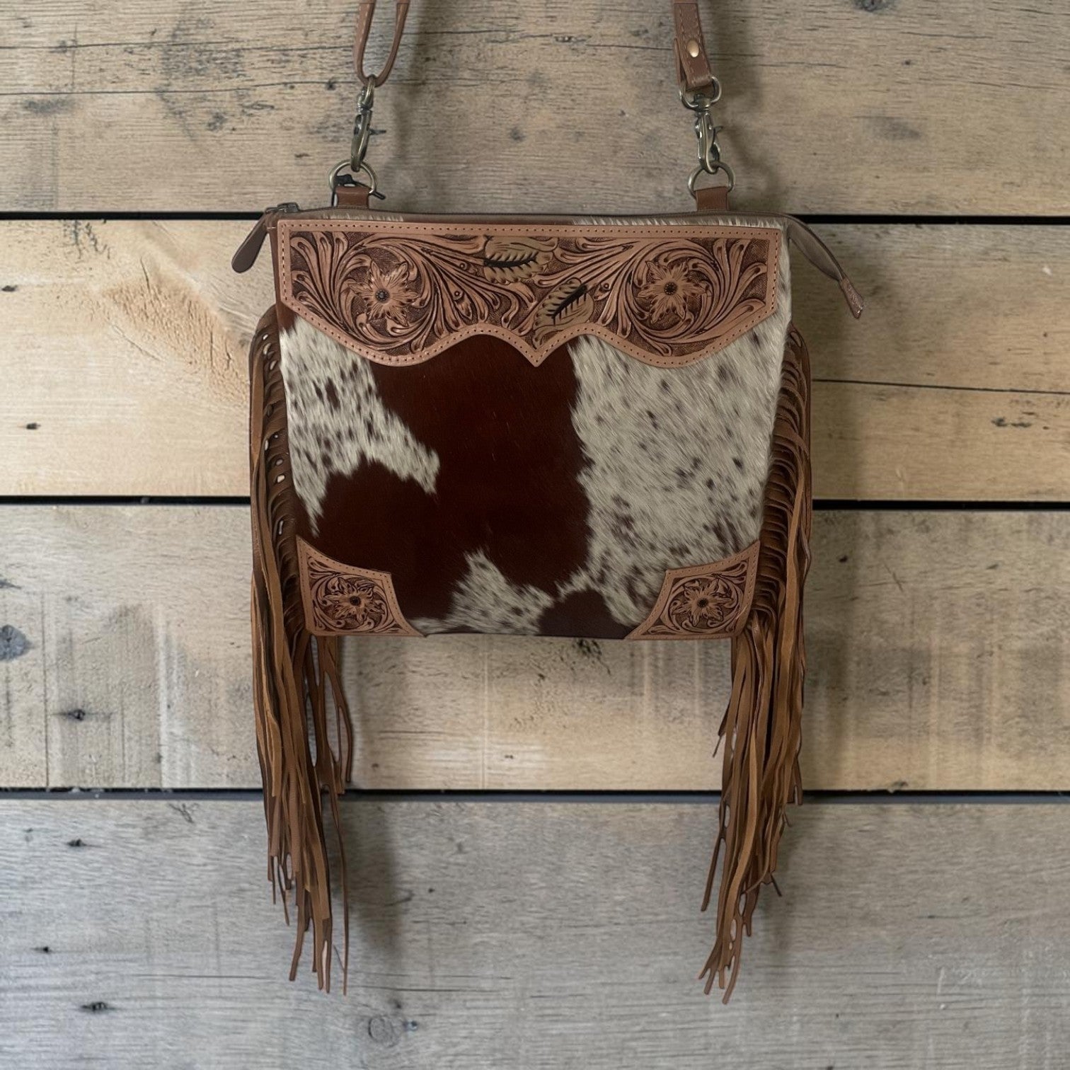 A western-style crossbody bag with a brown and white cowhide body, tooled leather trim, and long fringe, hanging against a rustic wood plank background.