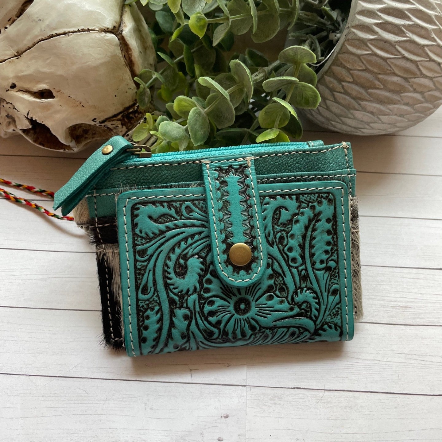 Turquoise leather wallet with intricate designs on a light wooden surface with plants and shells.