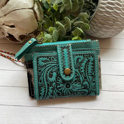Turquoise leather wallet with intricate designs on a light wooden surface with plants and shells.