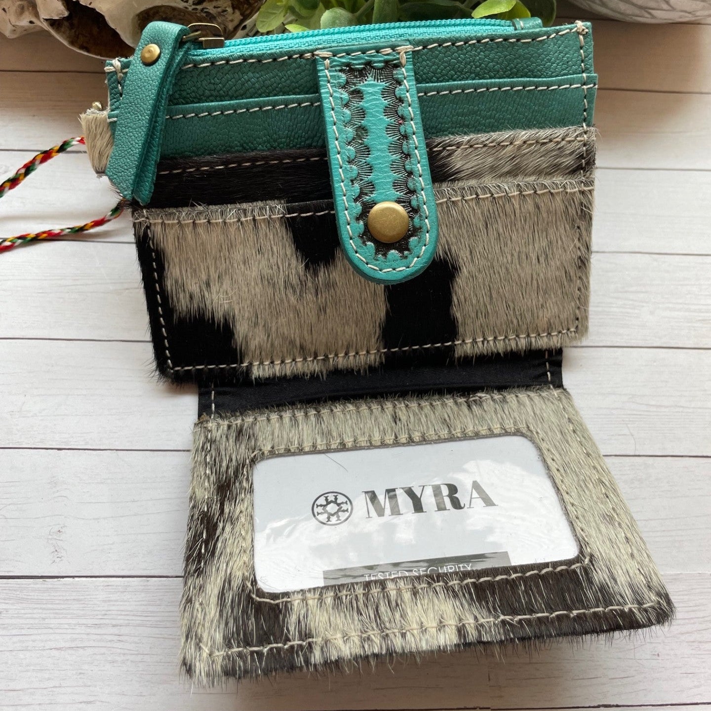 Teal and cowhide patterned wallet with 'MYRA' branding on a light wooden surface.
