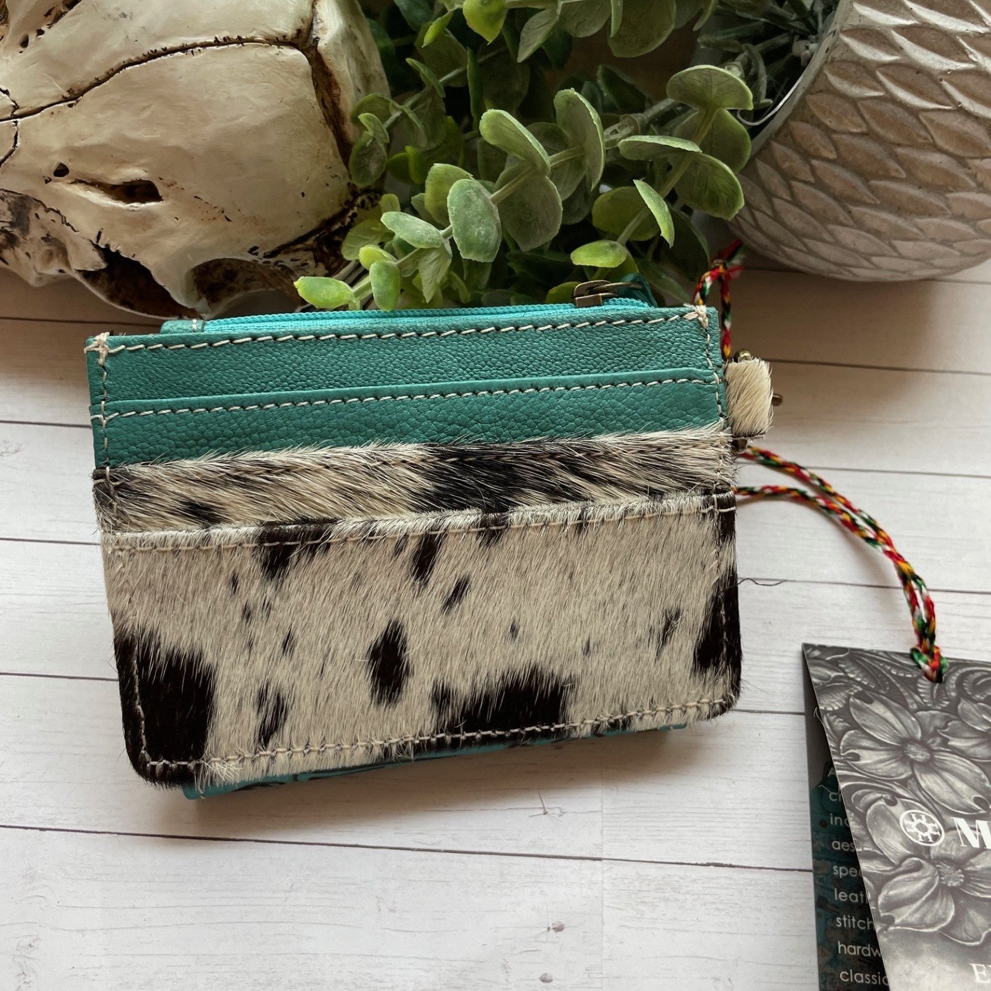 Cardholder with cowhide design and teal strip on a wooden surface with plants and skull.