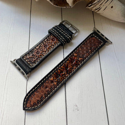 A brown tooled leather watch strap with floral embossing and white contrast stitching, displayed on a white wooden surface next to a decorative bone.