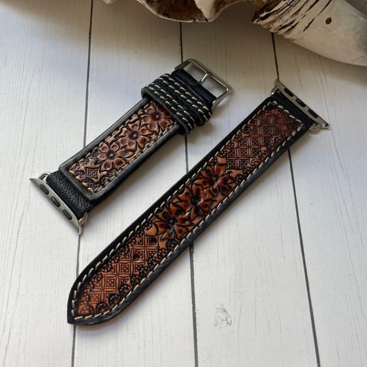A brown tooled leather watch strap with floral embossing and white contrast stitching, displayed on a white wooden surface next to a decorative bone.