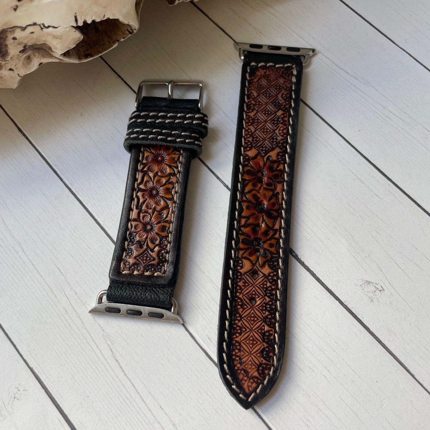 Two brown leather watch straps with floral tooled patterns and silver-toned hardware, displayed on a white wood plank surface next to a decorative skull.