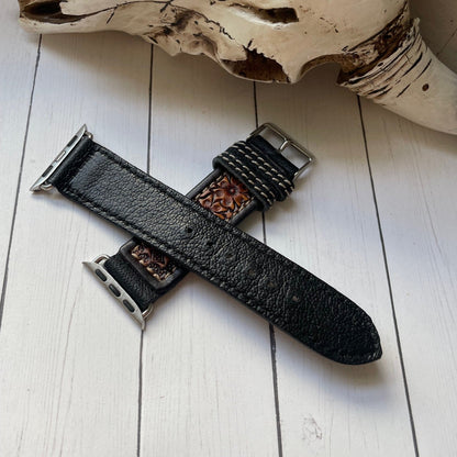 A black leather watch strap with silver-toned hardware and a brown patterned inner lining, laid on a white wood plank surface with a decorative animal skull in the background.