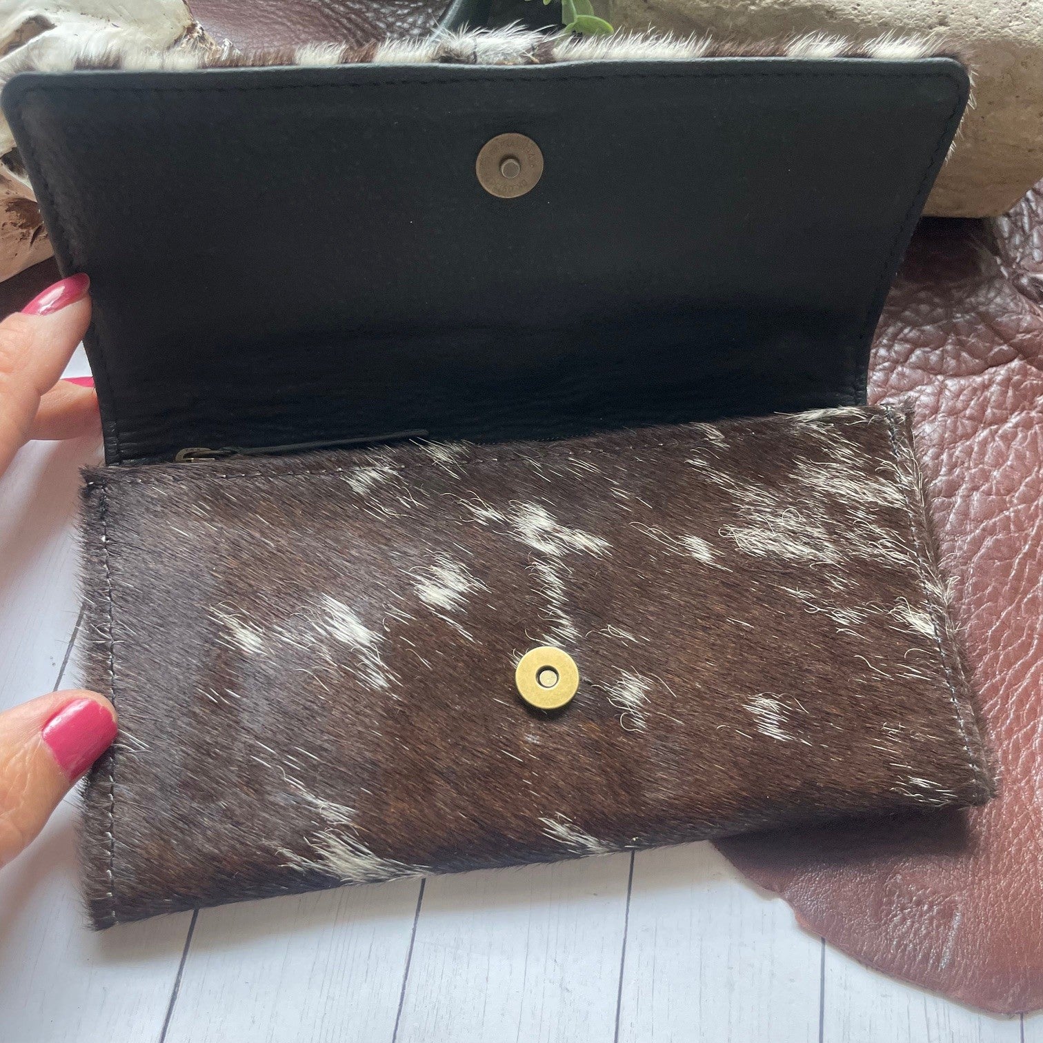 A brown and white cowhide wallet held open by a hand with pink nail polish, showing a black interior lining and a gold snap button closure.