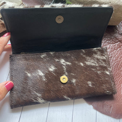 A brown and white cowhide wallet held open by a hand with pink nail polish, showing a black interior lining and a gold snap button closure.