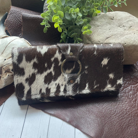 A cowhide wallet with a brown and white spotted pattern, featuring a circular metal ring detail on the front, displayed on a rustic surface with leather and a plant.
