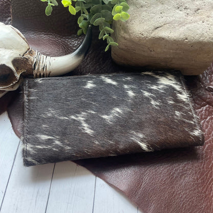A folded bison-print wallet with dark brown and white cowhide pattern, styled on a brown leather surface with a skull, green plant, and stone.