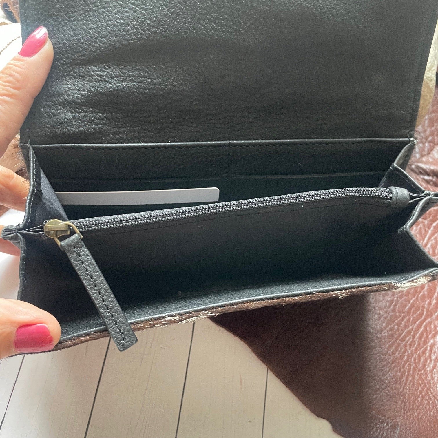 An open black leather wallet or clutch shown from the inside, with a hand holding the left edge. The wallet has a zippered compartment and card slots, photographed over a light wood floor and brown leather surface.